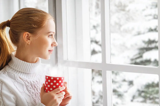 A woman in winter clothes holding a warm cup and looking through a window.